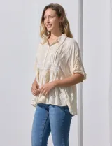 Blusa color crema de crepe con cuello a la base con solapa y escote en V con abertura central y botones. Delantera con detalles plisados y apliques en crochet. Manga corta con terminación fruncida. Ruedo en broderie.