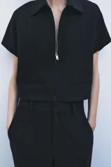 Chaqueta corta negra de cuello solapa y manga corta. Cierre frontal con cremallera metálica y bolsillos de ojal. Zara Woman Collection.
