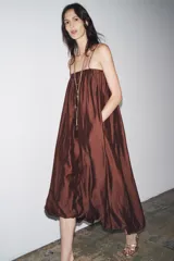 Vestido largo marrón de tejido satinado, con escote recto, tirantes finos extraíbles, bolsillos laterales ocultos y bajo asimétrico acabado en globo.