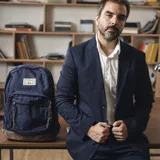 Mochila azul marino de tela Oxford con base de nobuk marrón, dos bolsillos frontales con cierre y tiradores de cuero, correas acolchadas y etiqueta de marca tejida en blanco y negro.