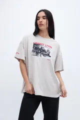 Remera de algodón color beige claro con efecto lavado, corte boxy y manga corta. Estampado frontal con una fotografía en blanco y negro de un local con el texto "LIBERTY LIVES" en rojo. Pequeño texto "CHARM" en la manga izquierda.