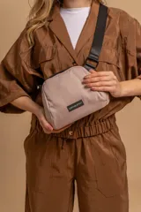 Bandolera pequeña de forma redondeada, color beige claro, con cierre superior negro y correa ancha ajustable de tela negra. Posee una etiqueta rectangular negra con texto blanco en el frente.