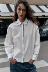 Camisa blanca de popelín con vivos negros a contraste en el cuello, el frente y los puños. Presenta un corte holgado y mangas largas con puños anchos.