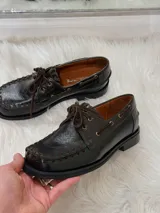 Zapato tipo mocasín de cuero color chocolate, con suela plana de goma negra, cordones al tono y costuras decorativas visibles en el borde superior.