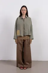 Camisa amplia de lino en tono verde desgastado, con textura visual y caída liviana. Destaca por su parche tejido a mano en contraste, que aporta un detalle artesanal y único.