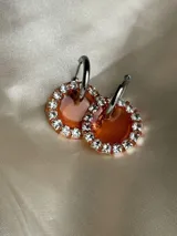 Aros colgantes con forma circular, hechos de resina color lila y borde de strass plateado. El aro es de acero quirúrgico plateado.