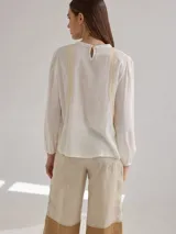 Camisa blanca de lino y viscosa, de corte holgado, con cuello redondo y mangas largas abullonadas. Presenta detalles de puntilla de algodón en el frente.