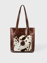 Bolso tipo tote de cuero color marrón con bolsillo externo de cuero con estampado animal print de vaca en blanco y marrón.