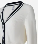 Cardigan corto de punto color crema con escote en V ribeteado en negro y blanco. Presenta cierre frontal de botones negros y puños con el mismo ribete a rayas. Las mangas son largas y ligeramente abullonadas.