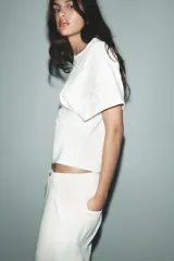 Camiseta blanca de algodón con cuello redondo y manga corta. Presenta bordados florales a tono en el lateral derecho del pecho y en la parte inferior izquierda.