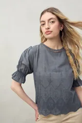 Blusa azul grisáceo de algodón con bordados frontales y terminaciones festoneadas. Tiene mangas cortas con frunces y cuello redondo con detalle de puntilla.
