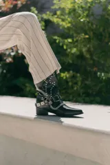 Bota de caña media color negro, estilo texana, con punta fina, tachas plateadas y apliques de hebillas metálicas con cadena.