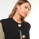 Chaleco de punto grueso color negro, sin mangas, con cuello redondo y cierre frontal con botones dorados.