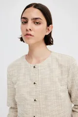 Saco corto de tweed color beige, con cuello redondo, cierre frontal con botones dorados y bolsillos delanteros decorativos.