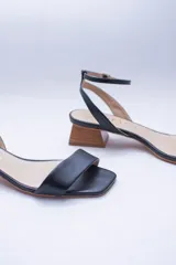 Sandalias de cuero negro con tira delgada sobre los dedos, pulsera al tobillo con hebilla dorada y taco geométrico color madera.