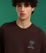 Remera marrón de corte relajado, confeccionada en algodón. Presenta cuello redondo, mangas cortas y estampa frontal y trasera de palmeras con la leyenda 'Work Hard'.