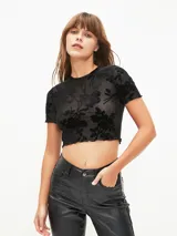 Top corto negro de manga corta, confeccionado en tejido de malla transparente con diseño de terciopelo floreado en relieve. Presenta cuello redondo y ruedo con terminación de ondas.