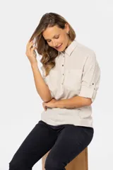 Camisa color beige de lino, de manga larga con botones metálicos personalizados y presilla en la manga. Tiene cuello clásico, cierre frontal parcial con botones y bolsillo de parche en el pecho.
