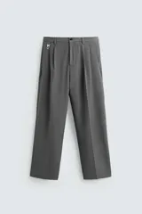 Pantalón gris de corte ancho con cintura elástica, pliegues frontales y trabilla con hebilla metálica. Incluye bolsillos delanteros y traseros de vivo.