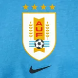 Remera blanca de algodón con cuello redondo y manga corta. Presenta un escudo estilizado de la Asociación Uruguaya de Fútbol (AUF) con cuatro estrellas doradas arriba y el logo de Nike en color dorado.