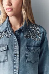 Camisa de denim celeste con apliques de piedras plateadas en los hombros, corte recto, cuello camisero, botones metálicos y bordado Rapsodia en la espalda.