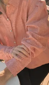 Camisa color rosa confeccionada en lino, de corte recto, con cuello clásico y mangas largas abullonadas con puños abotonados. Presenta tablitas verticales en el frente.