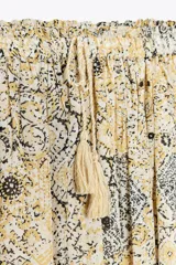 Falda larga color crema con estampado floral en tonos negro y dorado, cintura elástica con cordón ajustable y borlas decorativas.
