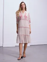 Vestido midi blanco con bordados florales azules en el escote, mangas y cintura. Tiene cuello en V, mangas 3/4 con botón y falda escalonada con detalles de ribetes azules.