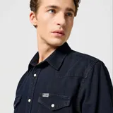 Camisa de denim azul oscuro con cuello americano, cierre con botones a presión blancos, dos bolsillos frontales con solapa y logo de la marca bordado en el bolsillo izquierdo.