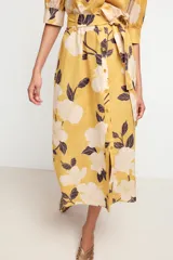 Vestido midi camisero color amarillo con estampado de flores blancas y hojas marrones. Tiene cuello solapa, mangas 3/4 con puños, cinturón de lazo ajustable y cierre frontal con botones.