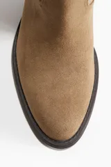 Botas de caña alta estilo cowboy, color marrón claro (camel/taupe), con acabado de gamuza o nobuck. Presentan costuras decorativas en la caña, punta de almendra y taco ancho y bajo de color oscuro.