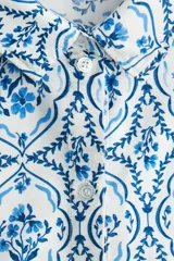Camisa de corte holgado con estampado de flores azules sobre fondo blanco, cuello clásico, botones frontales y mangas cortas. Confeccionada en sarga con canesú y pliegue en la espalda, hombros caídos y bajo redondeado con aberturas laterales.