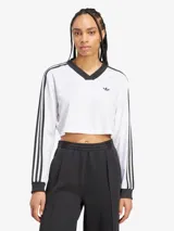 Top corto blanco de manga larga con cuello en V y ribete negro. Presenta las tres rayas características de Adidas en las mangas y el logo bordado en el pecho.