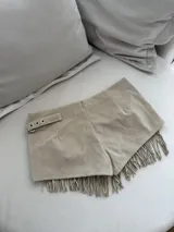 Minifalda color beige de gamuza sintética, con tiro bajo, corte irregular, tachas plateadas y flecos en el ruedo.
