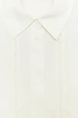 Camisa de lino color crudo, con cuello solapa y escote en pico. Tiene mangas por debajo del codo y detalle de entredós combinados a tono. El bajo es asimétrico y el cierre frontal es con botones.