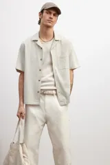 Camisa de manga corta color beige claro, confeccionada en tejido liviano de lino y algodón. Tiene cuello resort, tapeta lisa, bolsillo superior y bajo recto con aberturas laterales.