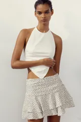 Falda pantalón corta color crudo con estampado de lunares negros, tiro medio, detalle de pliegues y volantes, y cierre lateral con cremallera.