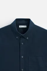 Camisa azul marino de corte regular fit, confeccionada en tejido de algodón Oxford. Tiene cuello abotonado, manga larga con puños abotonados, bolsillo de parche en el pecho y cierre frontal con botones.