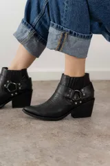 Botas cortas de cuero negro con puntera fina, caña baja con elásticos laterales y hebilla decorativa.