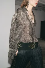 Camisa de viscosa con estampado paisley en blanco y negro. Cuello redondo con escote pico ajustable con cordones, mangas largas con elástico y volantes en los puños, bajo asimétrico y cierre frontal con botones forrados.