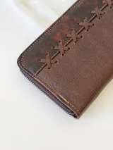 Billetera de cuero marrón con cierre metálico y diseño de cruces cosidas en el frente. Incluye monedero interno y espacio para billetes extendidos.