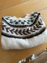 Sweater tejido a mano en lana mohair color crudo, con cuello redondo y mangas largas. Presenta un diseño con motivos geométricos en tonos marrón y verde en la parte superior del pecho.