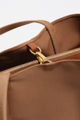 Bolso shopper grande de nylon negro, con estructura blanda, doble asa de mano y correa larga ajustable para llevar al hombro. Presenta un logo metálico con el texto "BIMBA Y LOLA" en la parte frontal inferior.