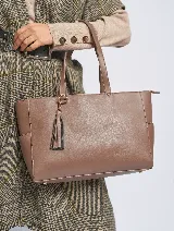 Cartera tote color marrón topo, de cuero sintético, con doble asa corta y cierre superior. Incluye un dije colgante con borla.