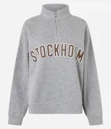Buzo gris de algodón sin felpa, modelo oversized, con cuello alto y cierre corto frontal. Presenta el texto "STOCKHOLM" estampado en el pecho con borde blanco y relleno marrón.