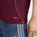 Camiseta de fútbol Adidas del AS Roma, modelo titular 25/26. Diseño color borgoña con detalles en naranja en cuello, mangas y hombros. Incluye logo de Adidas, escudo del club y tecnología Aeroready.