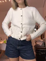 Cardigan de punto grueso color blanco roto, con botones oscuros y aplique floral de crochet en el lateral inferior derecho.