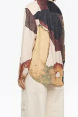 Camisa de corte recto con cuello camisero y botones frontales. La tela presenta un efecto arrugado o texturizado en tono beige claro, con grandes bloques de estampado abstracto en tonos tierra (marrón, ocre, verde oliva) que simulan manchas o texturas orgánicas. Las mangas son largas y una de ellas tiene un detalle en negro liso en el puño.