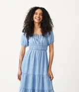 Vestido midi de jeans celeste, con escote cuadrado con lazo, mangas cortas abullonadas, cintura elastizada y falda escalonada con volados.