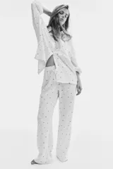 Pijama de dos piezas en muselina de algodón color crema con estampado de pequeñas figuras oscuras. Incluye una camisa de manga larga con cuello camisero, botones frontales y bolsillo de parche, y un pantalón recto con cintura elástica y cordón de ajuste.